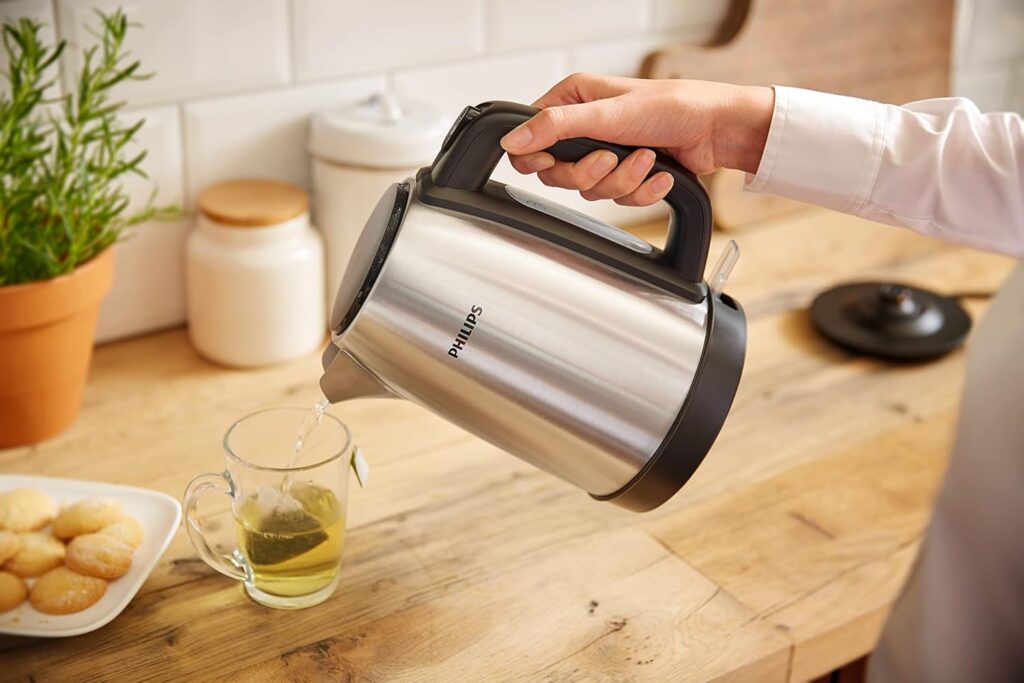 Person pouring hot water from a stainless steel Philips electric kettle HD9350 into a clear glass mug with a tea bag on a wooden kitchen countertop