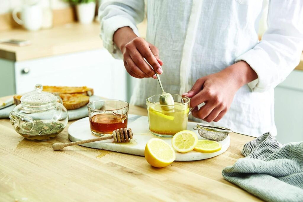 Person stirring a mug of hot tea on a wooden kitchen counter alongside a glass teapot, honey dipper, fresh lemon slices, and strainer.
