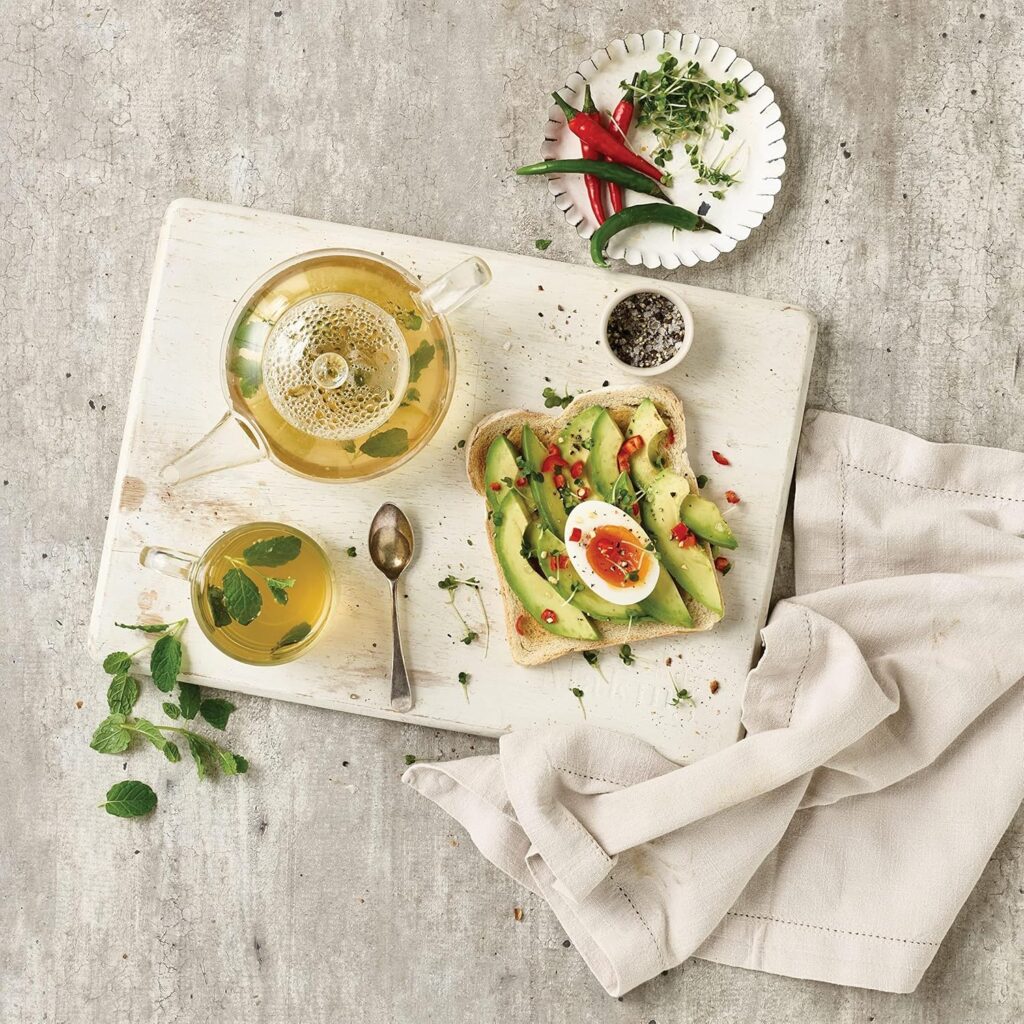 Top-down view of freshly brewed herbal mint tea in a glass teapot and teacup served with avocado egg toast on a wooden board.