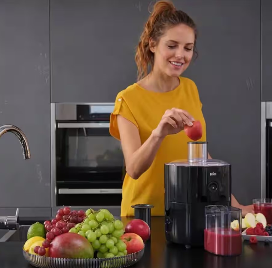 Smiling woman in a yellow top placing a slice of red apple into the chute of a black Braun SJ3100 FastXtract fruit juicer on a dark kitchen counter.