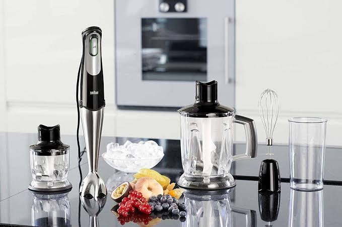 Braun MultiQuick 7 MQ7045 hand blender displayed on a kitchen countertop alongside its mini chopper, jug blender attachment, whisk, beaker, and fresh fruits with ice.