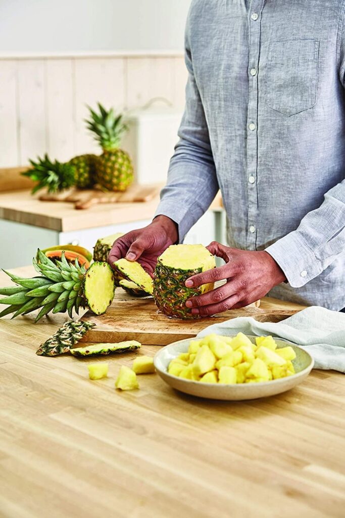 Person slicing a fresh pineapple on a wooden cutting board with cubed pineapple pieces in a bowl nearby.