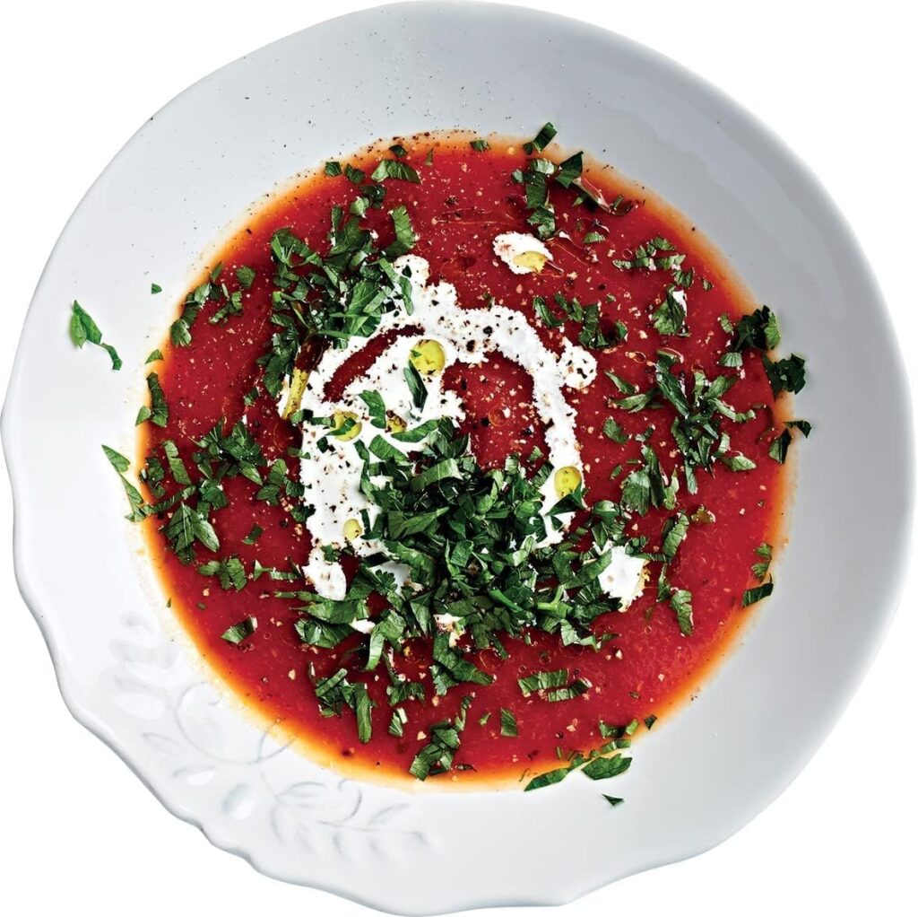 Top-down view of a white ceramic bowl containing rich red tomato soup topped with cream, olive oil drizzle, and chopped fresh herbs.