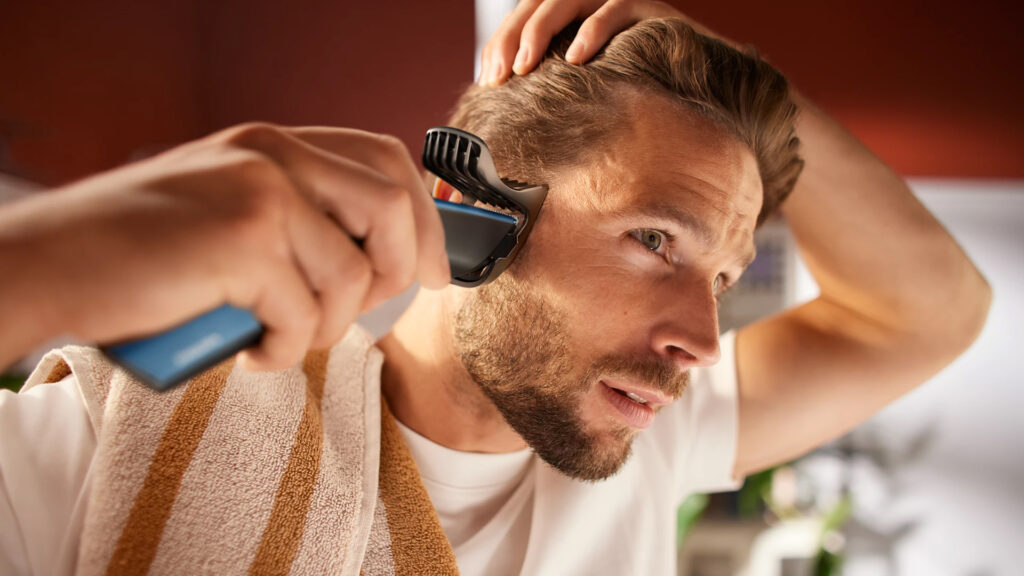 A man using the Philips Hair Clipper HC3505 to trim his hair near the temple in front of a mirror with a towel draped over his shoulder.
