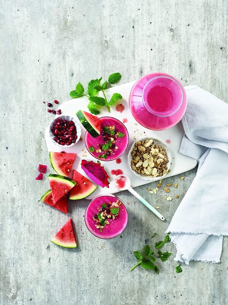 Top-down view of vibrant pink smoothie glasses and pitcher served on a white board with sliced watermelon, dragon fruit, pomegranate seeds, and granola.