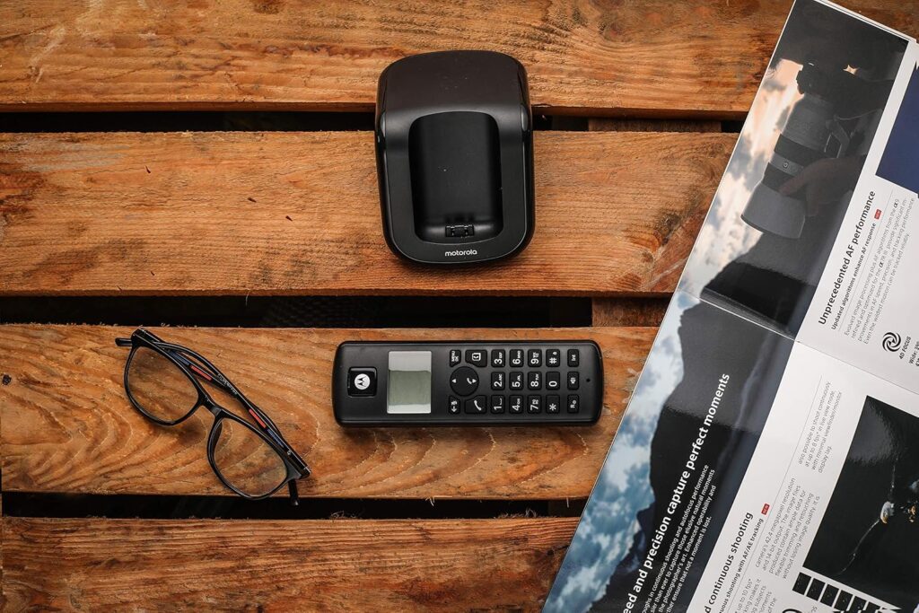 Motorola T201+ cordless phone handset next to its charging base on a wooden table, with glasses and a newspaper nearby